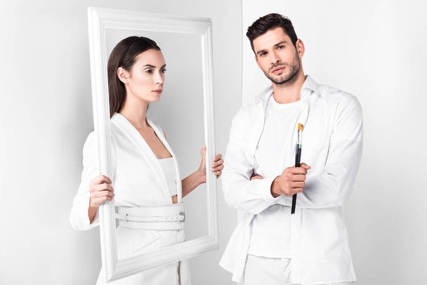 attractive adult woman holding frame near man with paintbrush on white background  - Foto, imagen