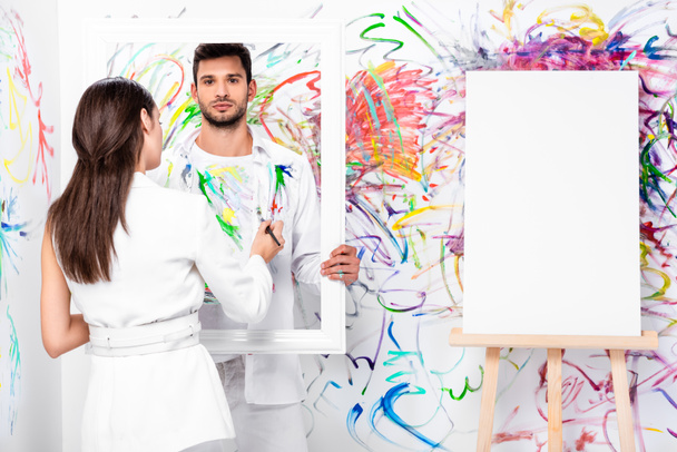 beautiful adult woman drawing on clothes while man holding frame near painted wall - Foto, imagen
