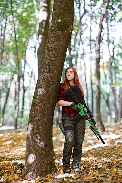 Soldat femme dans la forêt / Jeune femme avec fusil à la main se tient près d'un arbre dans la forêt d'automne
 - Photo, image