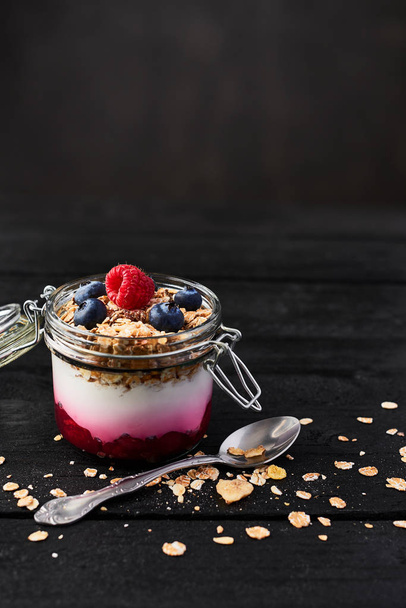 Healthy breakfast. Jar of healthy yogurt with raspberry sauce, oat and blueberries on black wooden table over dark concrete background with copy space. - Foto, Imagem