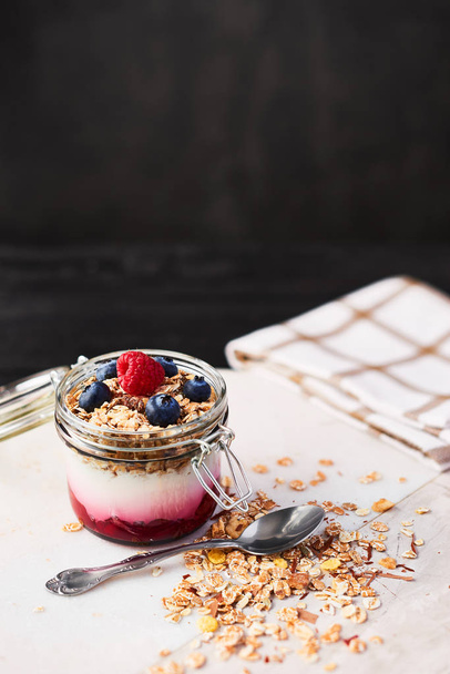 Healthy breakfast. Jar of healthy yogurt with raspberry sauce, oat and blueberries on white marble serving plate over dark concrete background with copy space. - Foto, imagen
