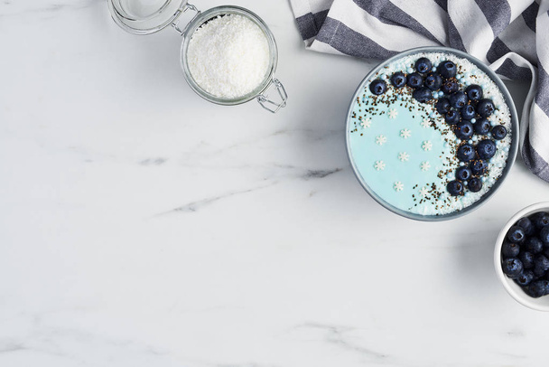 Top view of blue yogurt smoothie bowl made with blueberry, coconut flour, chia seeds and sugar pearls on white marble table with copy space. - Foto, Imagem