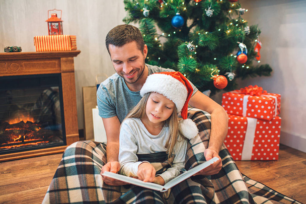 Father and daughter sitting on floor on blanket and reading book. They keeps it opened. Young man smiles. Girl is calm and peaceful. She reads book. They sit in room with fireplace and Christmas tree. - Photo, image
