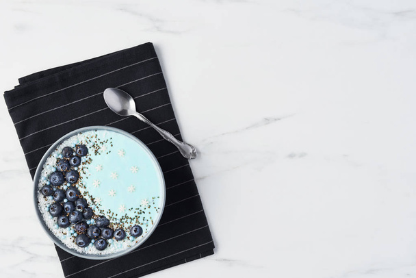 Top view of blue yogurt smoothie bowl made with blueberry, coconut flour, chia seeds and sugar pearls on white marble table with copy space. - Photo, Image