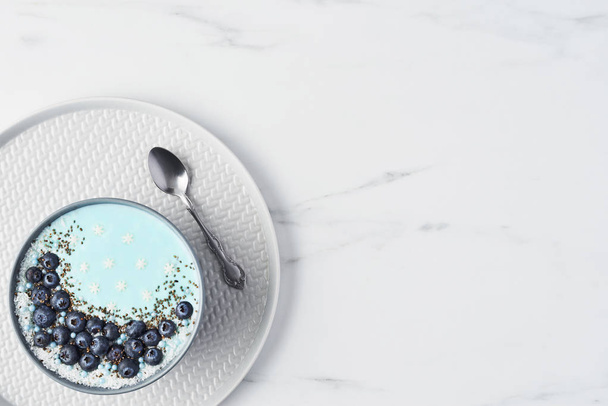 Top view of blue yogurt smoothie bowl made with blueberry, coconut flour, chia seeds and sugar pearls on white marble table with copy space. - Fotoğraf, Görsel