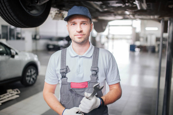 Portrait of young man in grey uniform standing and looking on camera. He smiles a bit. Guy wears uniform. He holds big wrench in hands. - Foto, Imagen