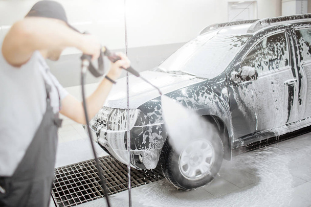 Another picture of man standing in room and washing cars wheel abd tire. He usies flexible hose with water for that. He is concentrated on process. - 写真・画像