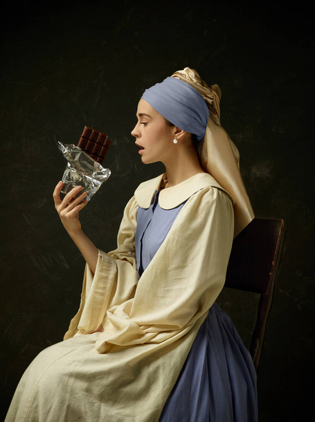 Medieval woman in historical costume wearing corset dress and bonnet. Beautiful peasant girl wearing thrush costume with sweet chocolate bar over dark studio - Foto, afbeelding
