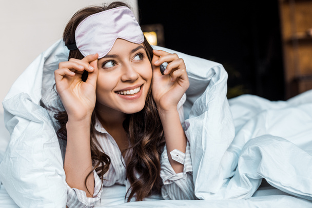 веселая девушка в маске для сна лежит под одеялом в постели
 - Фото, изображение