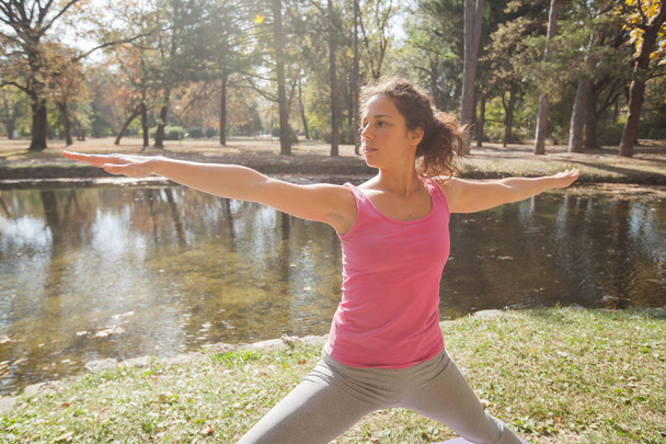 湖の近くの公園で戦士ポーズでヨガの練習を行う若い女性.美しい朝の秋の日。健康的な動機. - 写真・画像