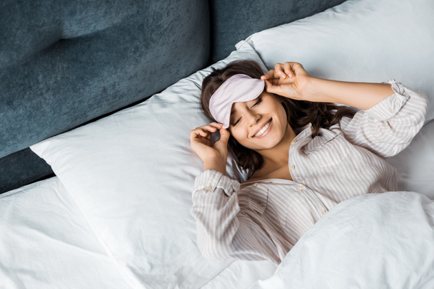 attraktives, glückliches Mädchen mit schlafender Augenmaske entspannt sich morgens im Bett  - Foto, Bild