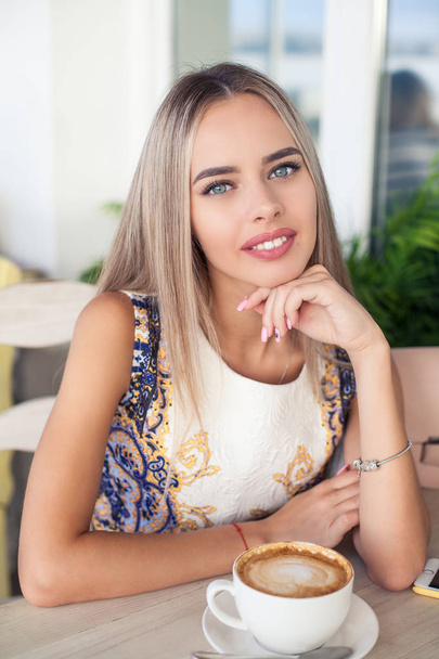 Young beautiful blonde with blue eyes is sitting in a cafe. A young woman holds a cappuccino mug in her hands. Portrait of a young happy woman. Fashion. Makeup. Summer. - Foto, afbeelding