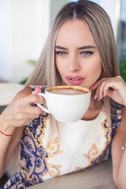 Young beautiful blonde with blue eyes is sitting in a cafe. A young woman holds a cappuccino mug in her hands. Portrait of a young happy woman. Fashion. Makeup. Summer. - Foto, afbeelding