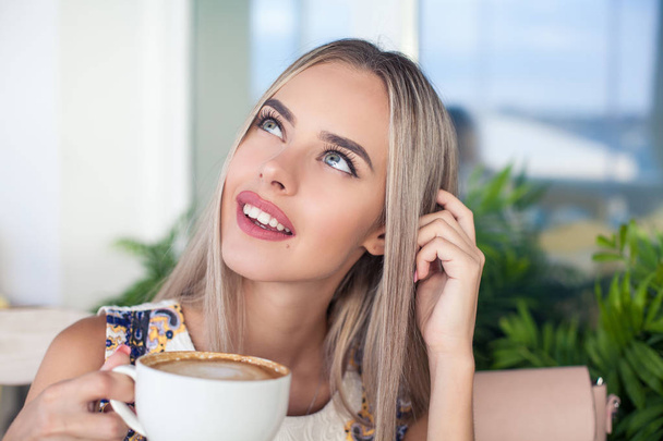 Young beautiful blonde with blue eyes is sitting in a cafe. A young woman holds a cappuccino mug in her hands. Portrait of a young happy woman. Fashion. Makeup. Summer. - Photo, Image