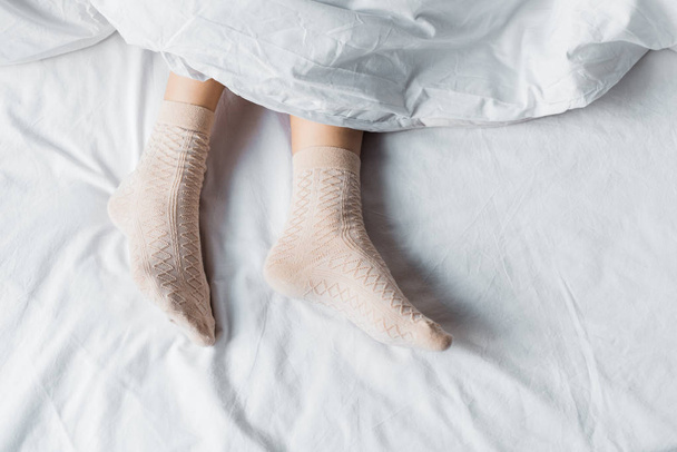 top partial view of girl in socks lying under white blanket in bed - Photo, Image