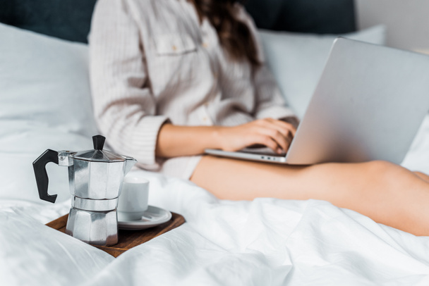 selective focus of moka pot with coffee cup and woman with laptop in bed - Photo, image