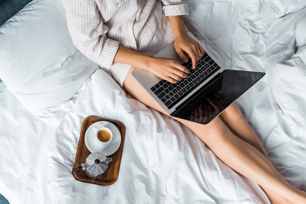 cropped view of woman with coffee cup and moka pot using laptop in bed in the morning - Foto, afbeelding