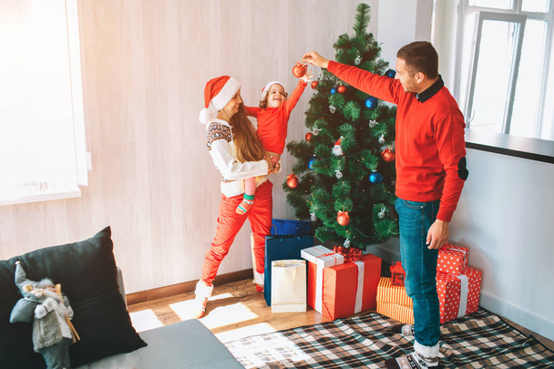 Merry Christmas and Happy New Year. Beautiful and bright picture of young family standing at Christmas tree. Man holds red toy and smiles. Child reaches to it with interest. Woman holds kid. - Photo, image