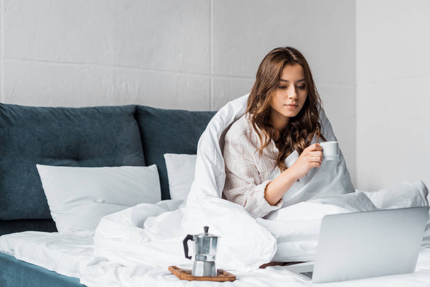beautiful woman with coffee looking at laptop in bed in the morning - Фото, изображение