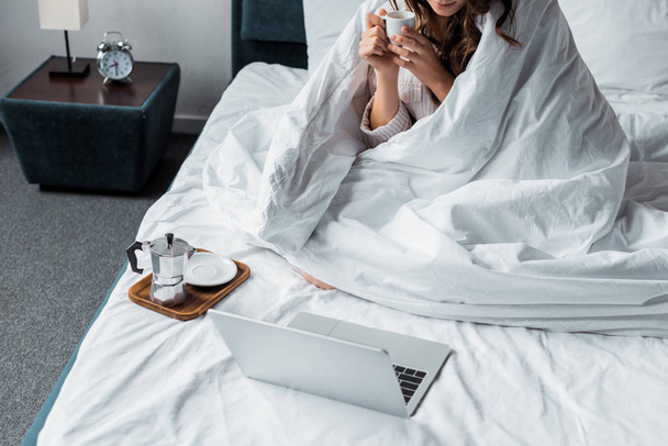 Ausgeschnittene Ansicht einer Frau mit Kaffeetasse, die auf Laptop im Bett schaut - Foto, Bild