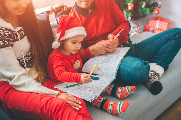 Merry Christmas and Happy New Year. Cut view of parents sitting on sofa with their child. Kid holds colouring and pencils on it. Girl draw with it. Young man holds some pencils too. - Photo, Image