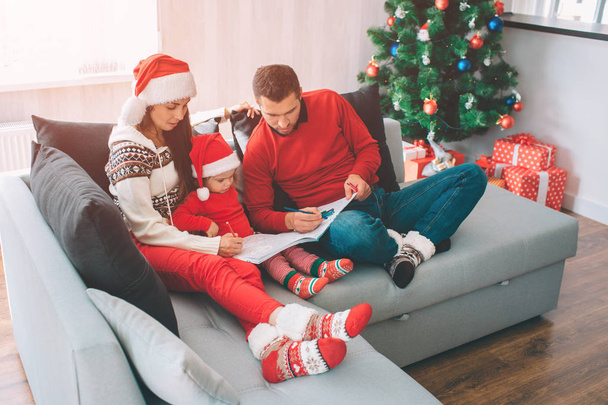 Merry Christmas and Happy New Year. Picture of adults drawing with pencils in colouring book. They are concentrated, calm and peaceful. Small girl look at that. She wears red clothes and hat. - Фото, зображення