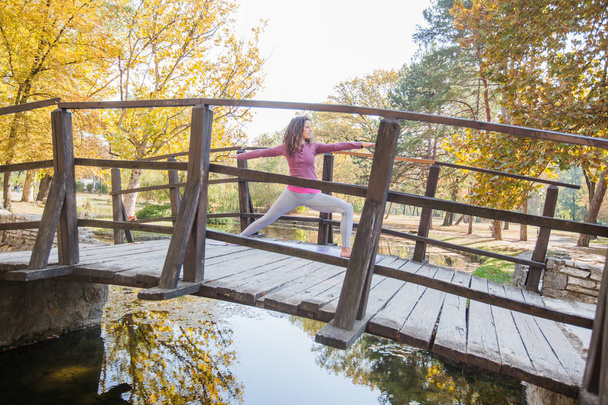 Young Woman Performing Yoga Exercise In Warrior Pose On Wooden Bridge At Park. Morning Autumn Day. Healthy Motivation. - Photo, Image