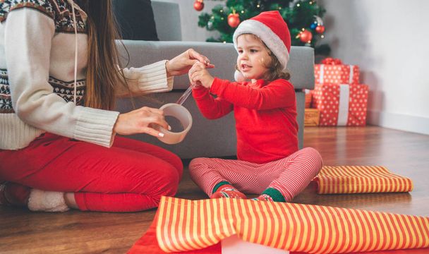 Merry Christmas and Happy New Year. Girl and young woman sit on floor. Child reaches and touches tape. Woman holds the rest of it. Child is excited. They prepare presents for beloved. - Photo, image