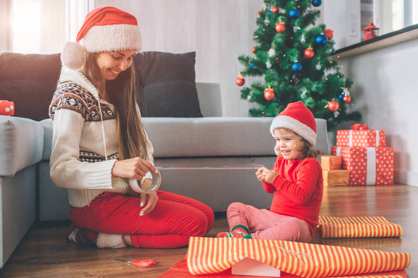 Merry Christmas and Happy New Year Positive and playful young woman and girl sit on floor. They smile and laugh. Child holds part of tape while woman has rest . They wear hats. Girls prepare gifts. - Foto, imagen