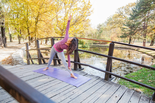 Flexibele jonge vrouw Yoga oefening Triangle Pose wilt uitvoeren op de houten brug bij stadspark. Sportieve Girl Training buiten. - Foto, afbeelding