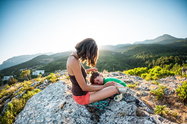 疲れた少年は、母親の足にあります。女性は、彼女の息子と旅行です。山の頂上に登るに疲れます。Mom は、山の頂上に座っている子を抱擁します。赤ちゃんが眠っています。. - 写真・画像