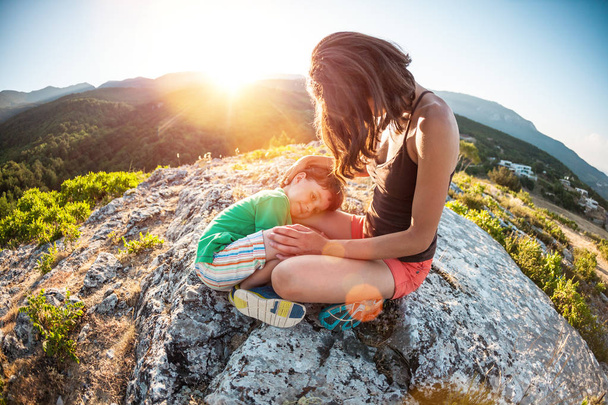 El niño cansado yace sobre los pies de su madre. Una mujer viaja con su hijo. Escalada agotadora a la cima de la montaña. Mamá abraza al niño, sentado en la cima de la montaña. El bebé está durmiendo
. - Foto, Imagen
