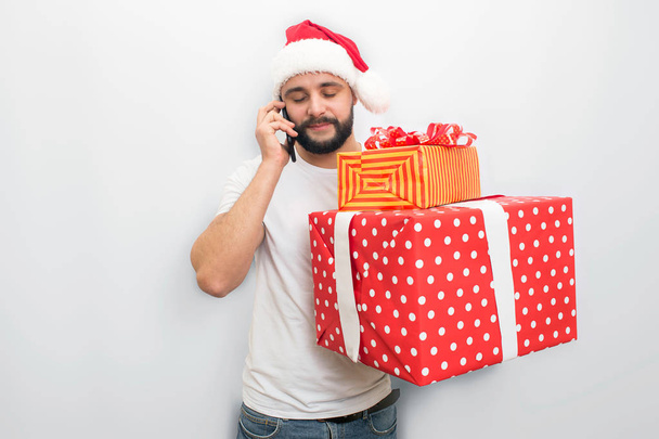 Busy young man stands and talks on phone. He holds bog red box with ribbons and present inside it. Guy looks down. Isolated on white background. - Φωτογραφία, εικόνα