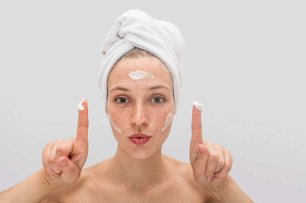 Professional model stands and shows her fingers with cream. Also there are some lotion on her forehead and cheeks. She has white towel around her hair. Isolated on grey background. - Foto, imagen