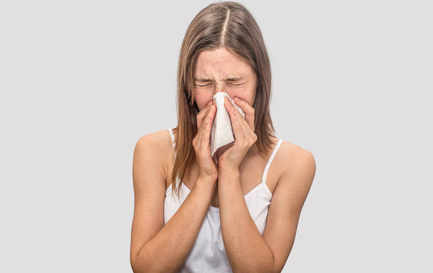 Sick and illyoung woman sneezing in dry napkin. She keeps eyes closed. Model suffering. Isolated on grey background. - Photo, image