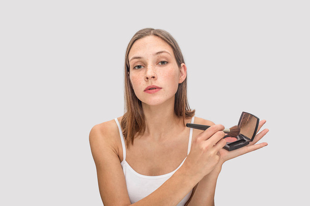 Young freckled model poses on camera. She holds pallete of eye shadows and use brush for that. Model looks on camera. She is cool. Isolated on grey background. - 写真・画像