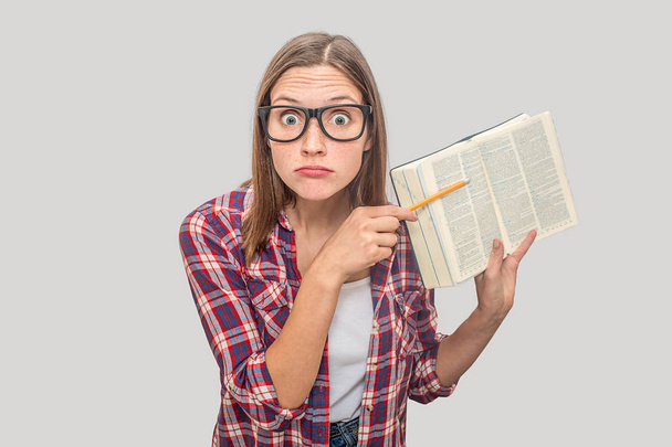 Weird and strange female geek keeps book wide opened and shows its pages on camera. She points on text with pencil. Young woman looks weird. Isolated on grey background. - Foto, Imagem