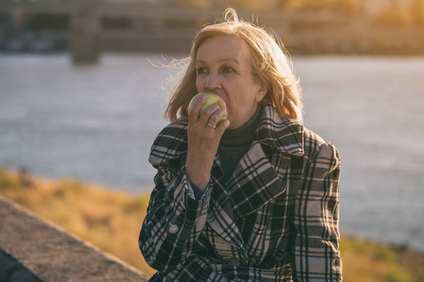 Ανώτερος γυναίκα απολαμβάνει διατροφικές apple ενώ κάθεται δίπλα στο ποτάμι. Πιο ήπια εικόνα. - Φωτογραφία, εικόνα