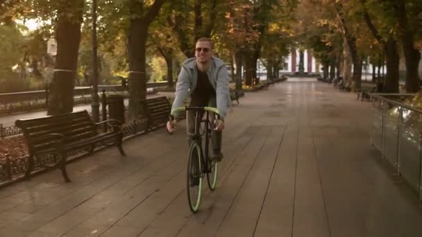 Front footage of a young smiling man in sunglasses cycling a bicycle in the morning park or boulevard. Slow motion of young man riding a trekking bike. Summer, autumn empty city park - Footage, Video