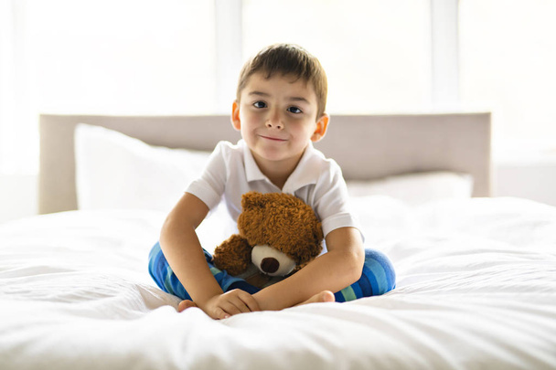 Uma criança senta-se na cama com um ursinho de pelúcia
 - Foto, Imagem