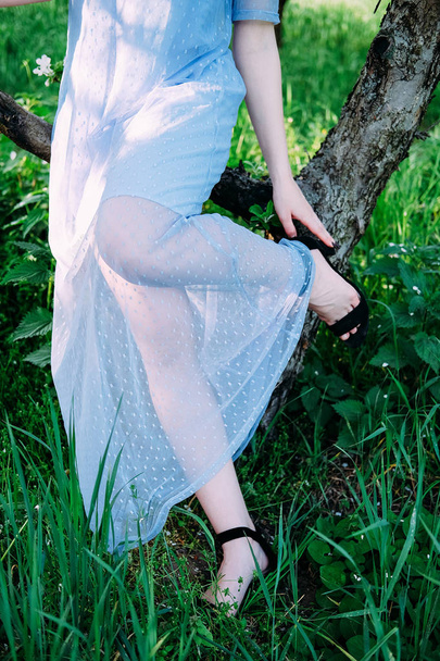 Calm and pretty young woman in the garden wearing transparent blue dress fixing up her shoes - Photo, image