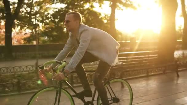 Side view of a young smiling man in sunglasses cycling a bicycle in the morning park or boulevard. Slow motion of young man riding a trekking bike. Summer, autumn empty city park - Filmmaterial, Video