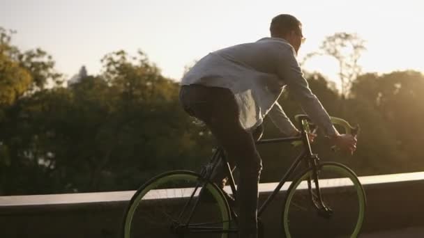 Happy young smiling man in sunglasses cycling a bicycle in the morning park or boulevard . Slow motion of young man riding a trekking bike, wind blowing and waves his shirt. Summer, autumn empty city - Filmmaterial, Video