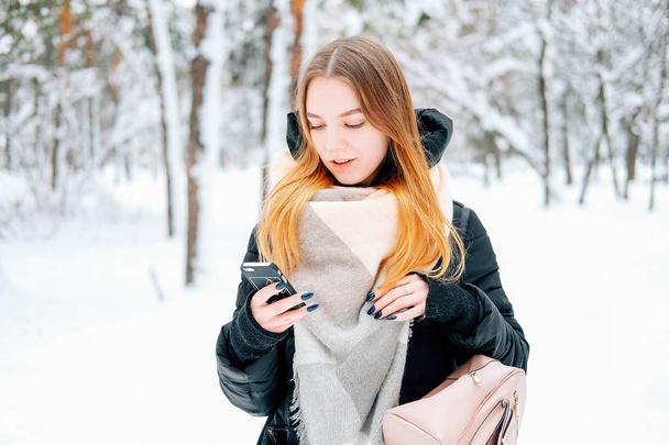 Aantrekkelijke blonde jonge volwassen vrouw lopen door winter Boshoogtepunt van sneeuw weearintg casual outfit van zwarte parka, jeans, roze leren laarzen en rugzak en beige sjaal en texting aan vrienden - Foto, afbeelding