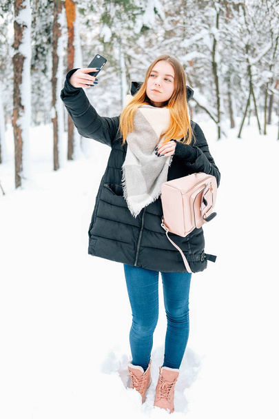Attractive blond young adult woman walking through winter forest full of snow weearintg casual outfit of black parka, jeans, pink leather boots and backpack and beige shawl and texting to friends - Photo, Image
