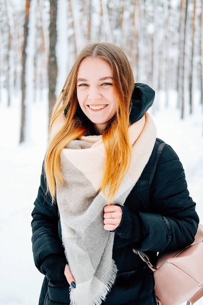 Attractive blond young adult woman walking through winter forest full of snow wearing casual outfit of black parka, jeans, pink leather boots and backpack and beige shawl - Photo, Image