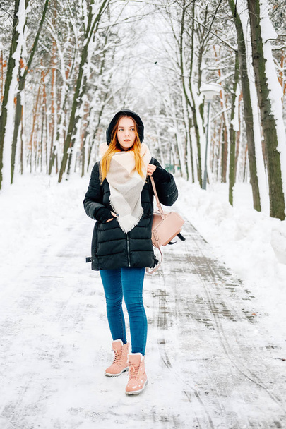 Aantrekkelijke blonde jonge volwassen vrouw lopen door winter Boshoogtepunt van sneeuw dragen casual outfit van zwarte parka, jeans, roze leren laarzen en rugzak en beige sjaal - Foto, afbeelding