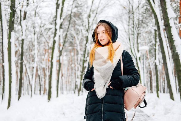 Attractive blond young adult woman walking through winter forest full of snow wearing casual outfit of black parka, jeans, pink leather boots and backpack and beige shawl - Photo, Image