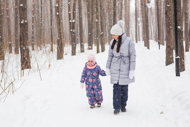Family, winter and nature concept - Young cheerful mother with cute little daughter walking in the snow park - Photo, Image