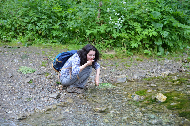 Mujer viajera lavándose la cara y las manos con agua del arroyo en el bosque. Chica se sienta con la mochila
 - Foto, Imagen